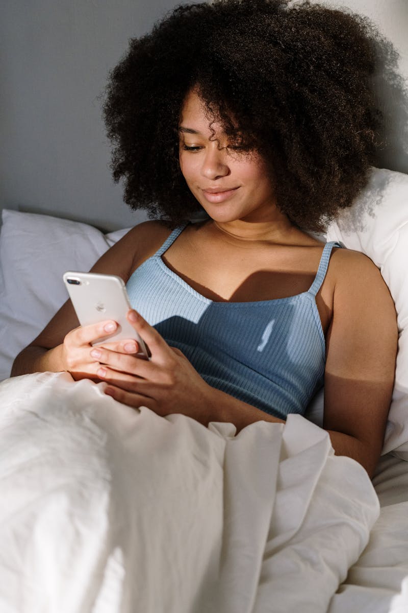 African American woman enjoying a cozy morning in bed, using a smartphone.